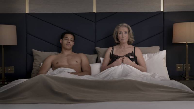 A shirtless young man sits under the sheets in a neat hotel bed. Next to him an older woman in a negligee sits upright, visibly tense.