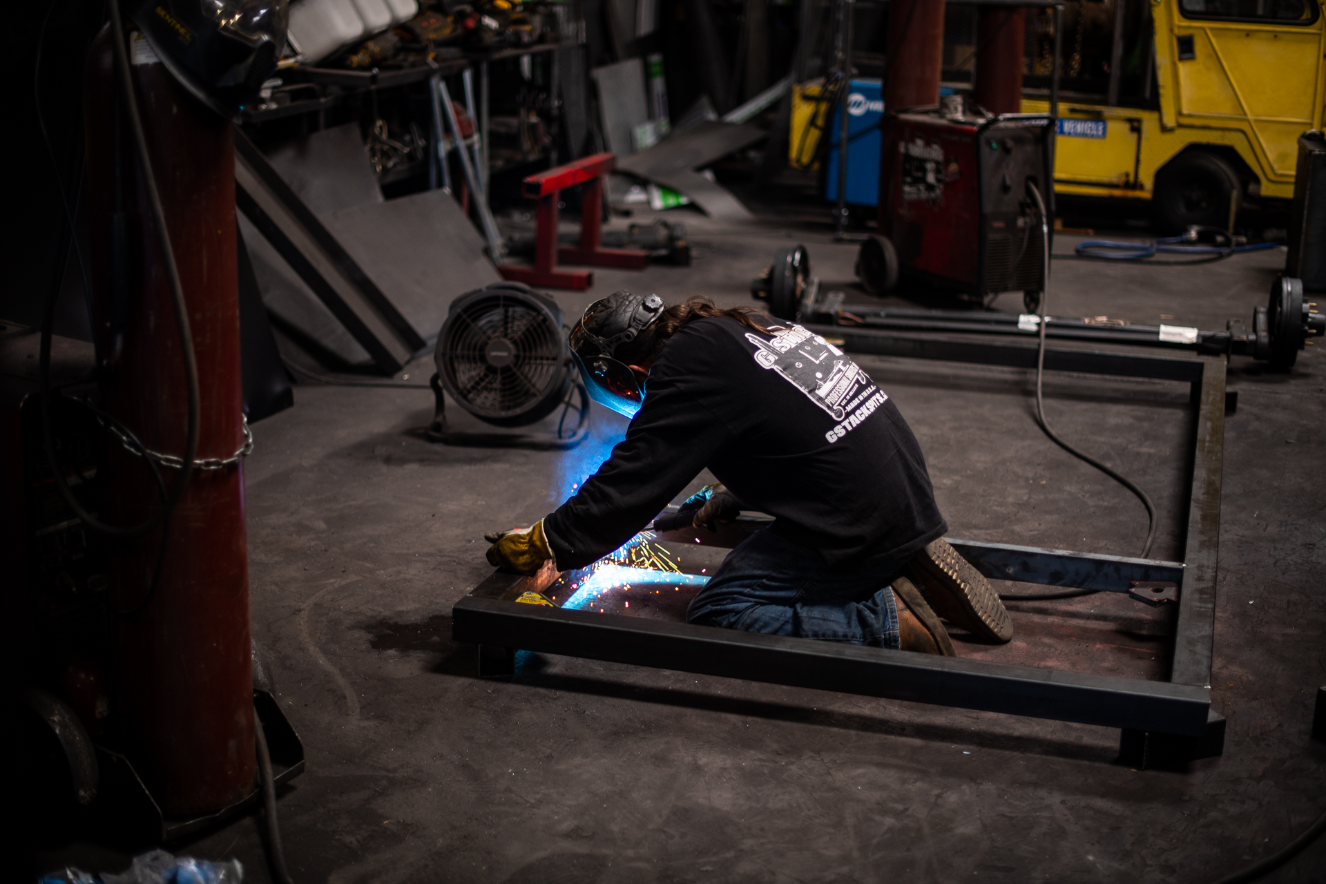 A man kneels on the ground welding something, as blue sparks fly.