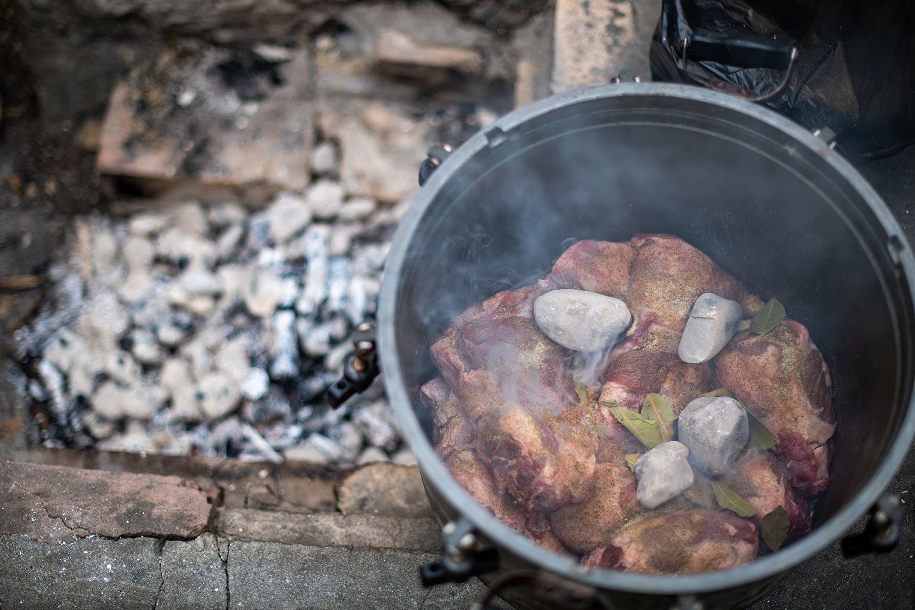 Overhead view of a pot filled with chunks of raw meat and rocks with wisps of smoke rising up.