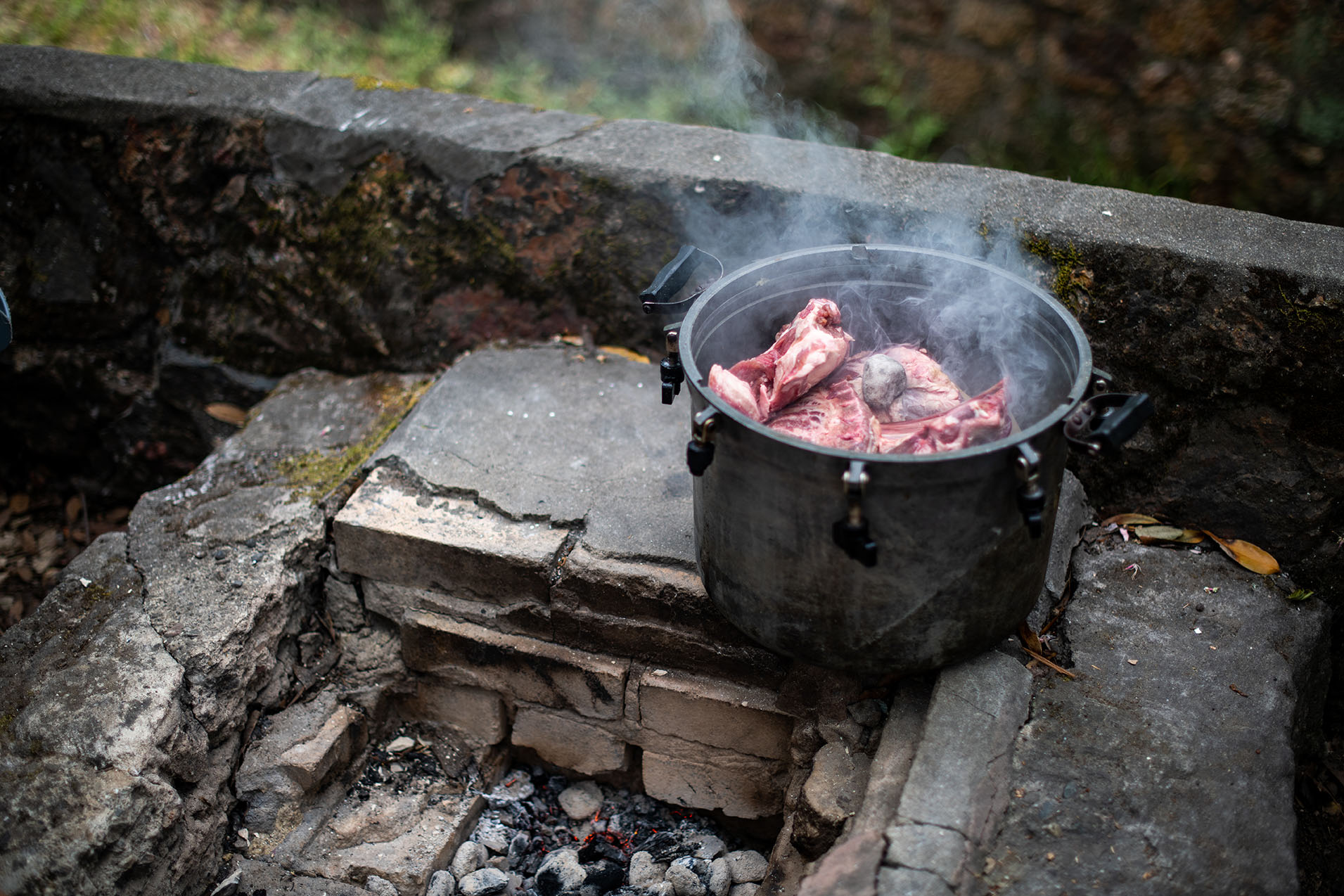 A metal pot filled with chunks of raw meat sits on an outdoor grill area with wisps of smoke rising up.