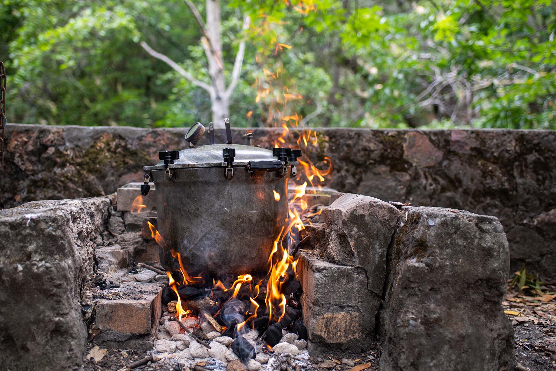 An old-fashioned pressure cooker heated over a fire outdoors.