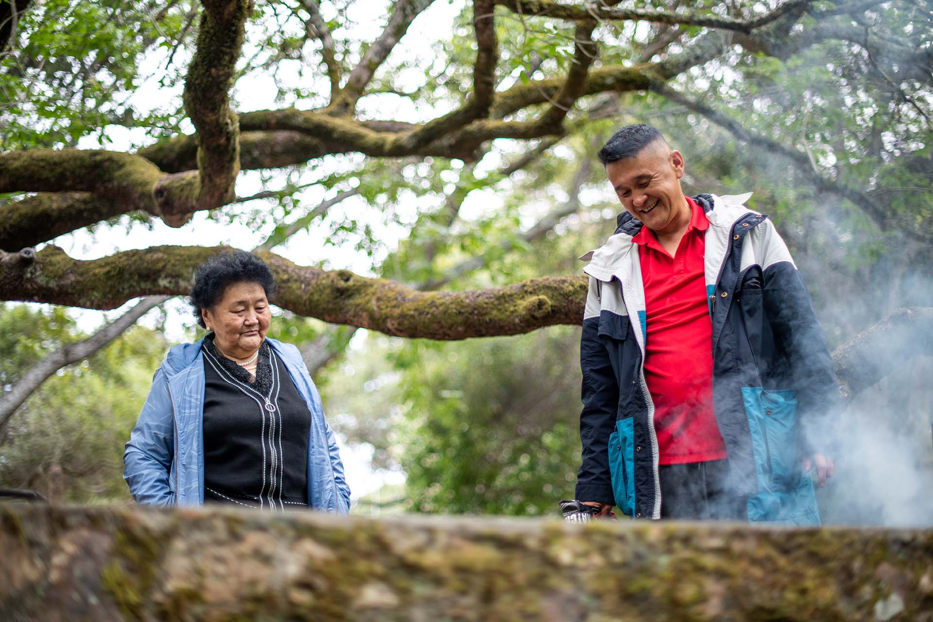 An older woman and a man smile, looking down toward the ground in a wooded area.