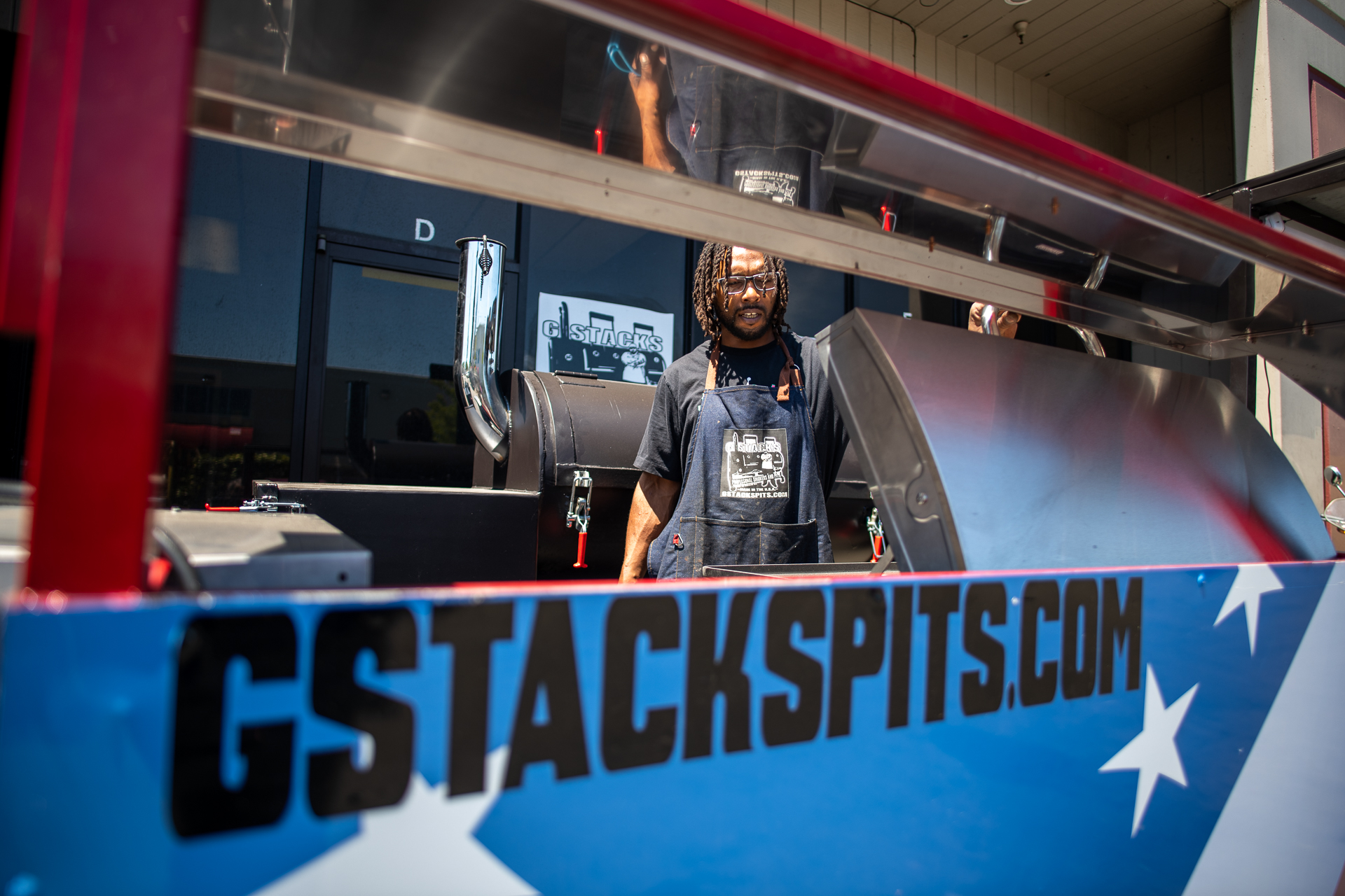 A man seen through an opening in a a barbecue smoker emblazoned with the website "GStackspits.com"
