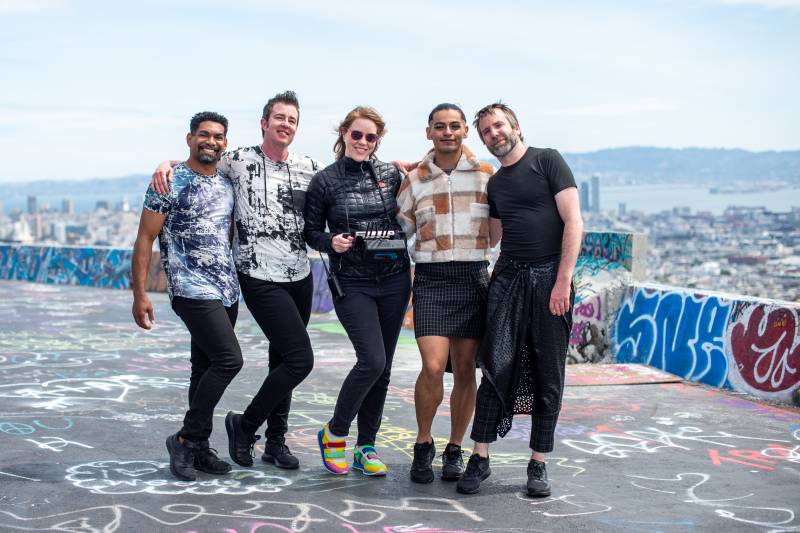 Four members of Sean Dorsey Dance are smiling and posing with filmmaker Lindsay Gauthier at the top of Twin Peaks with San Francisco's skyline behind them