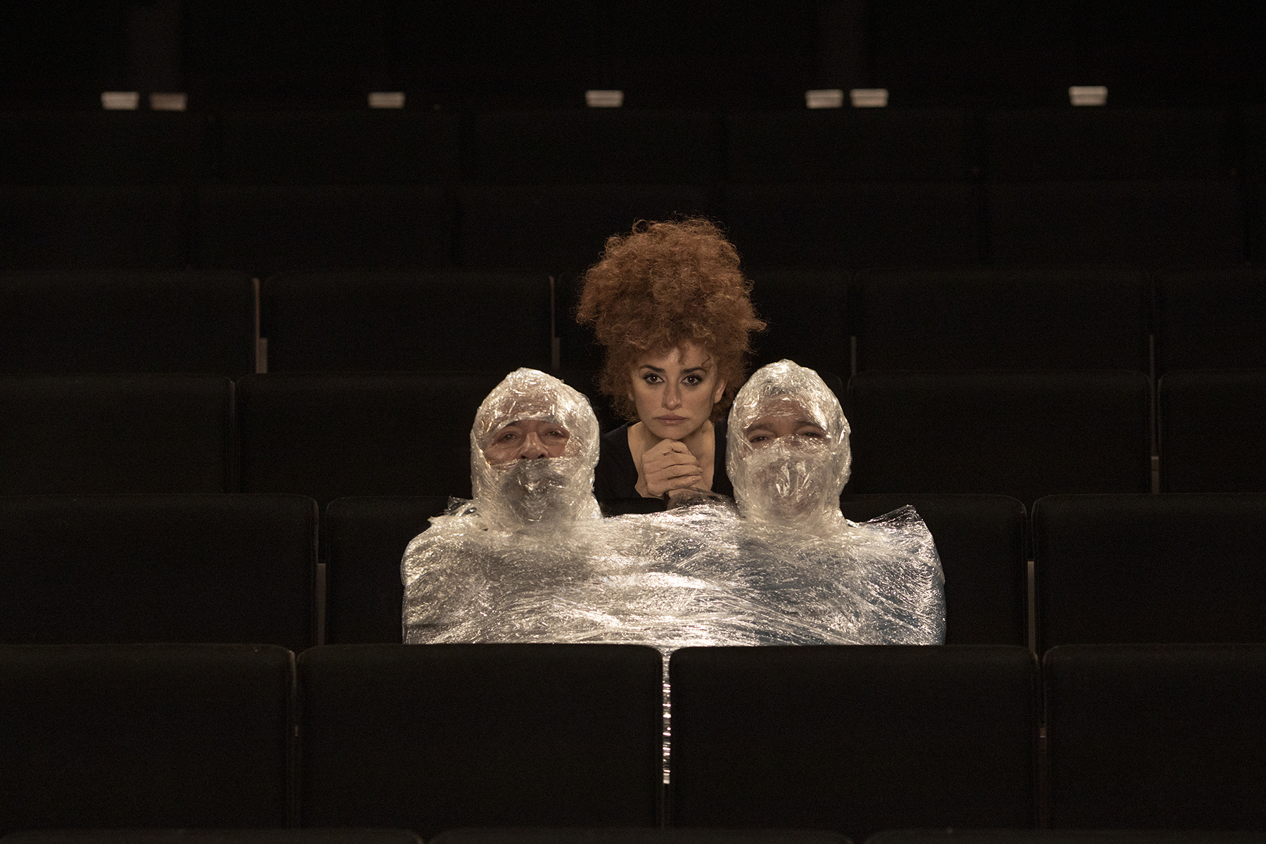 Two men wrapped in plastic like mummies, woman with curly hair between them in dark auditorium seats