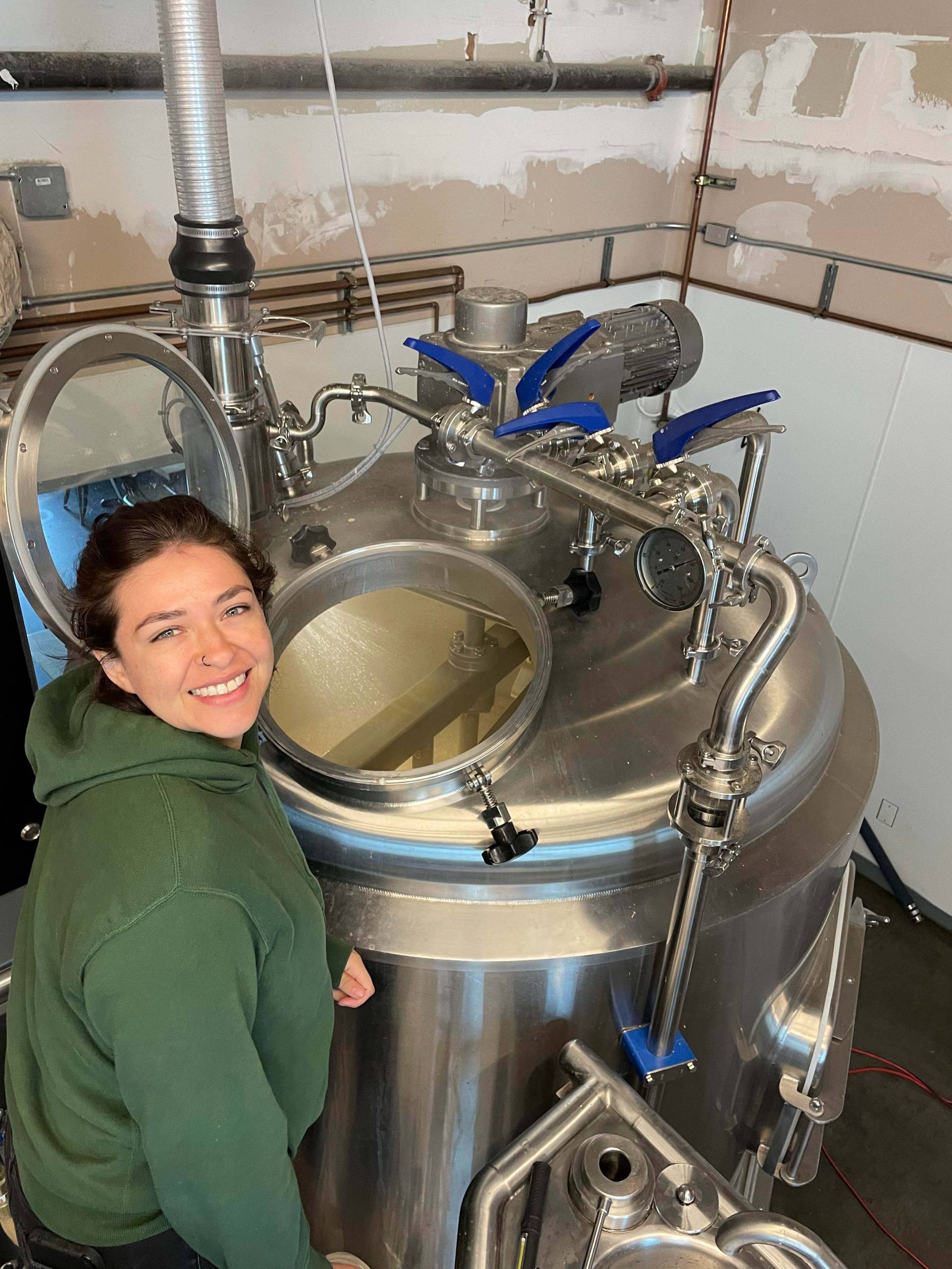 Woman in green sweatshirt stands in front of a large metal kettle for brewing beer.