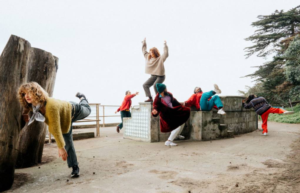 Six dancers in colorful clothes dance on and around large cement block in outside setting