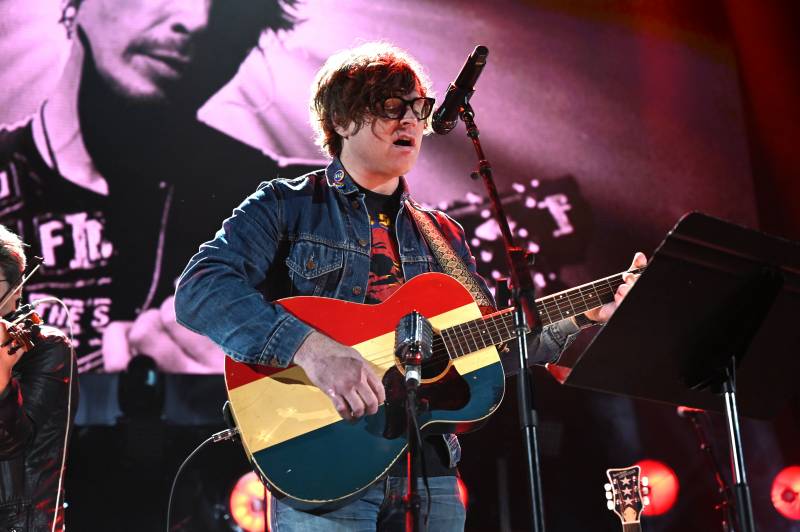 A man with scruffy hair and heavy-rimmed glasses performs on stage, playing an acoustic guitar.