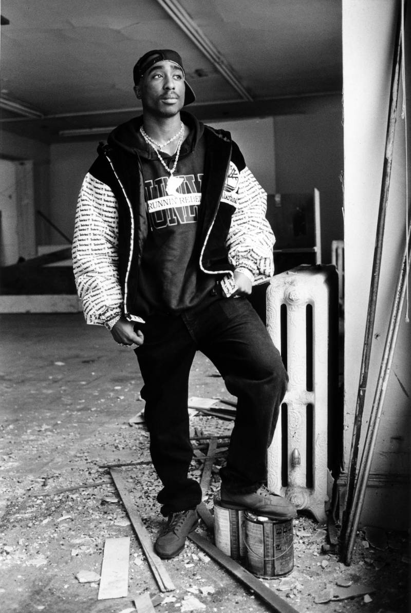A young Black man, the rapper Tupac Shakur, stands in a warehouse looking out the window in a black-and-white photo.