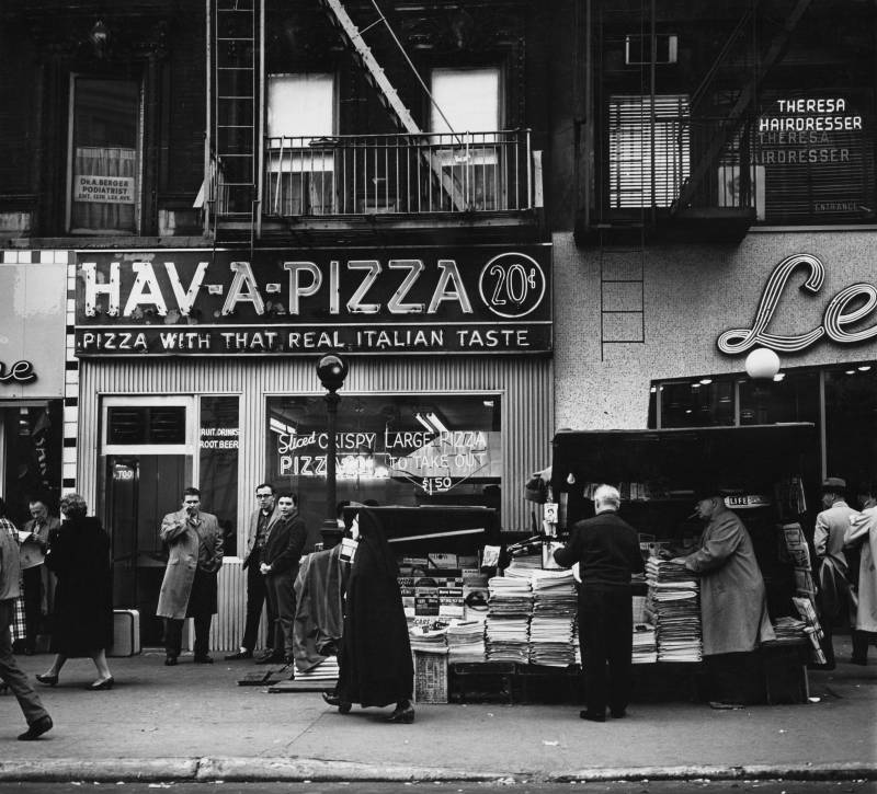 A black and white image shows a restaurant named Hav-A-Pizza with customers standing outside next to a busy news stand. A neon sign in the restaurant window advertises 20¢ slices.