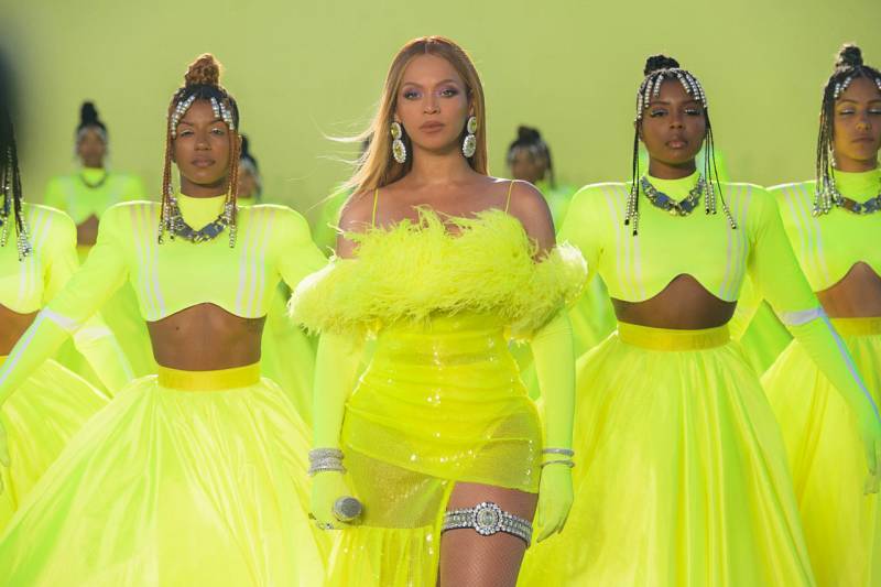 Beyoncé stands on stage in a luminous sheer green ensemble with feather bustier and high cut leg, diamond-encrusted garter on show. Behind her stand a formation of three strong-looking Black women wearing luminous green flowing skirts and tops.
