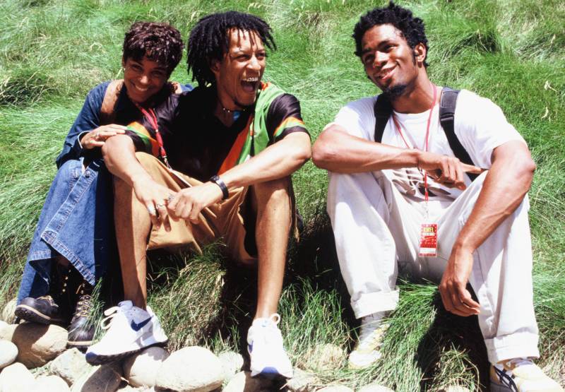 A hip-hop trio, one woman and two men, laugh while seated in the grass.