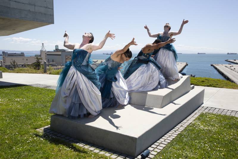 Four dancers in blue and white gowns pose on a concrete sculpture resembling a bed on a grassy lawn situated near the San Francisco Bay