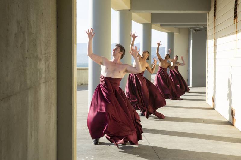 Four dancers in magenta gowns perform modern dance choreography against pillars at a cliff overlooking the Pacific ocean in San Francisco, CA