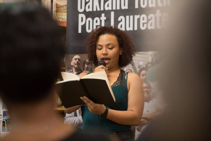A young woman in a tank top reads from a book in a crowded bookstore.