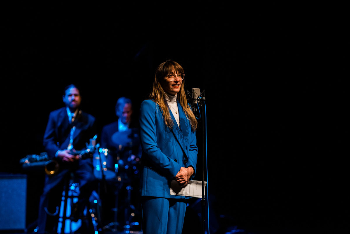 Woman in blue suit stands at mic in front of band