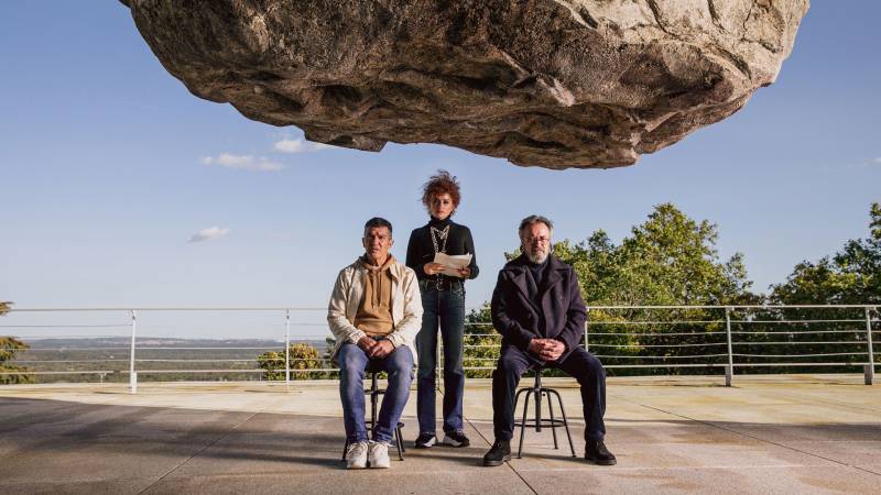 Two men sit on chairs, a woman stands between them, a giant rock is suspended above their heads