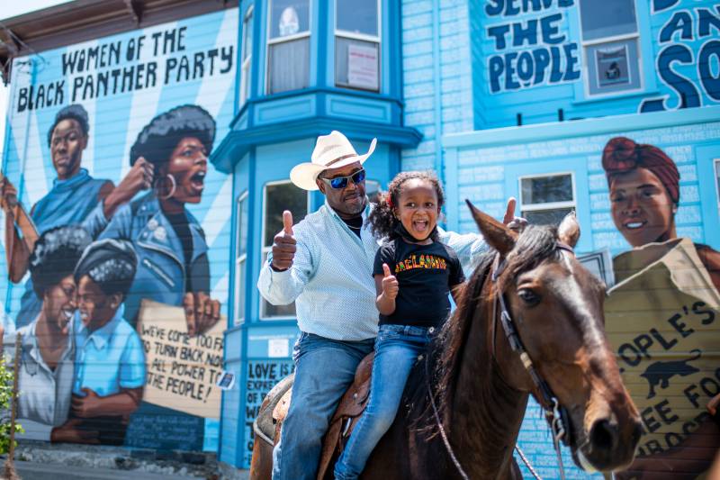 a young Black girl and an older Black man give thumbs ups while riding a horse in front of a mural celebrating the Black Panther Party