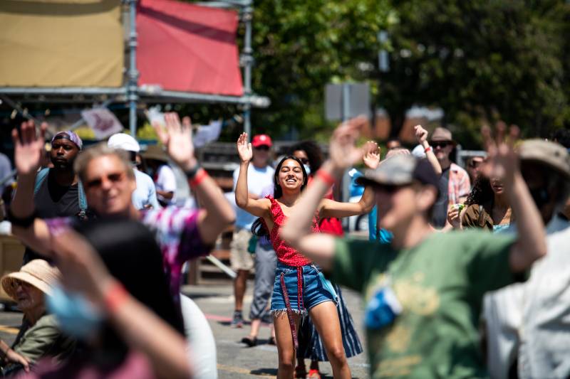 A group of people in bright clothes cheer and dance in the street during a block party to celebrate Juneteenth.
