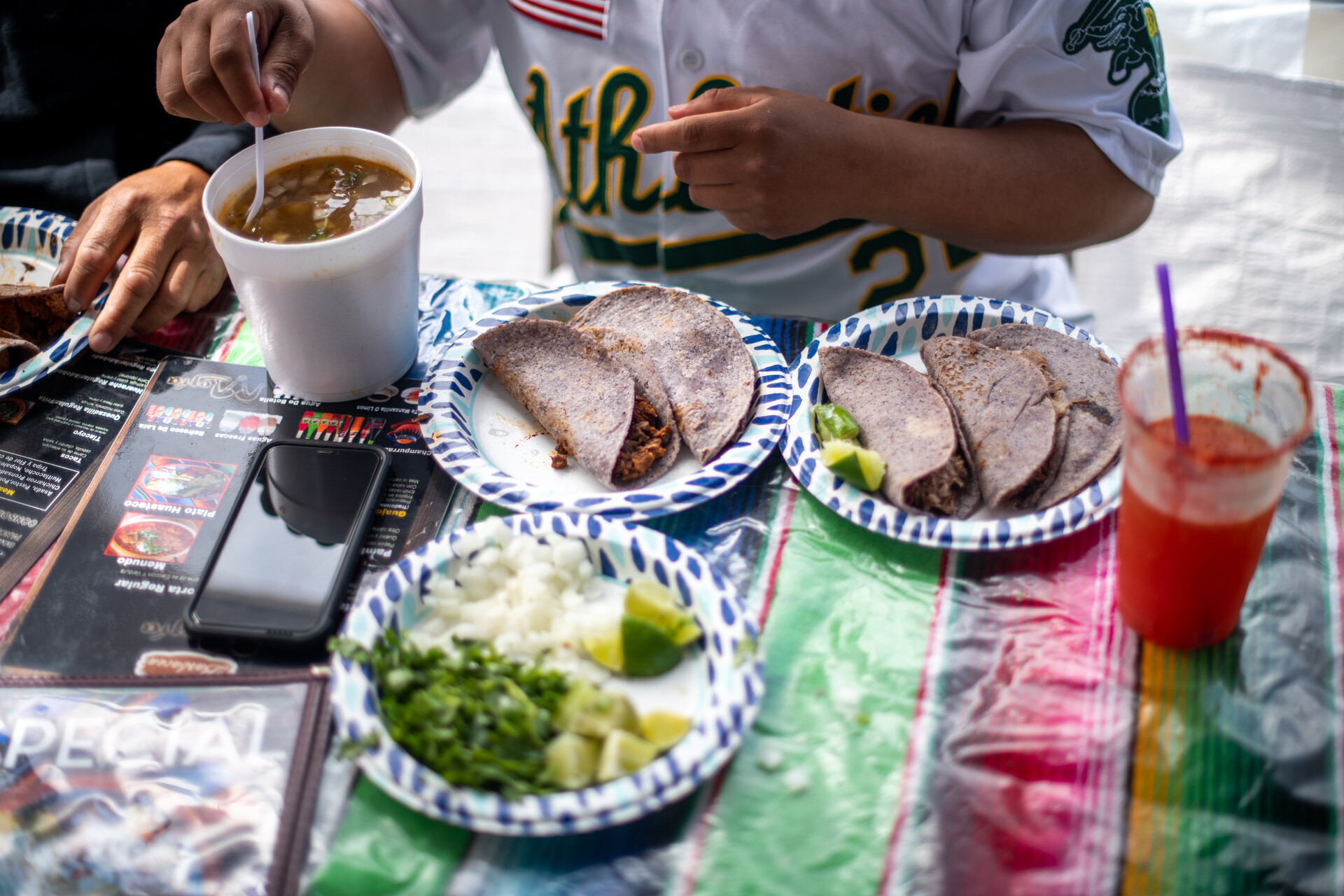 A diner in an Oakland A's jersey sits in front of a spread of blue corn tortilla tacos and all of their accompaniments, including a plate of onions, lime and cilantro.