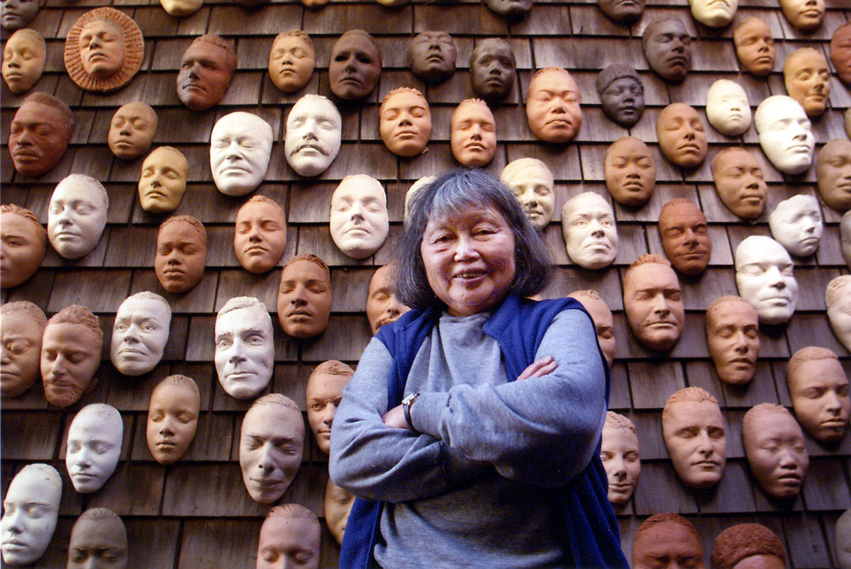 Older woman with crossed arms in front of shingled wall covered in clay masks