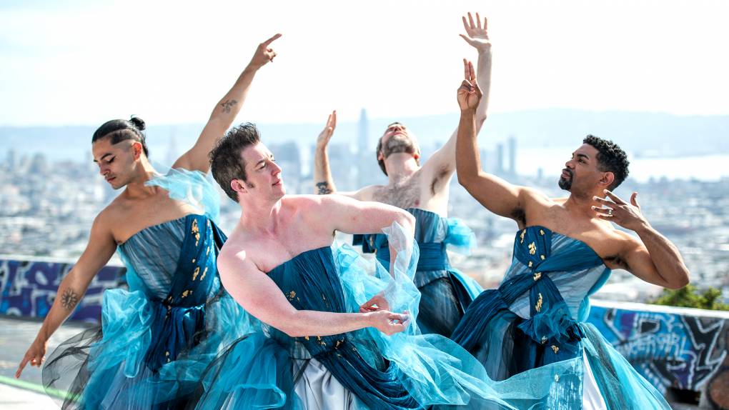 Four dancers in blue gowns are posing against a wide shot of San Franciso's skyline