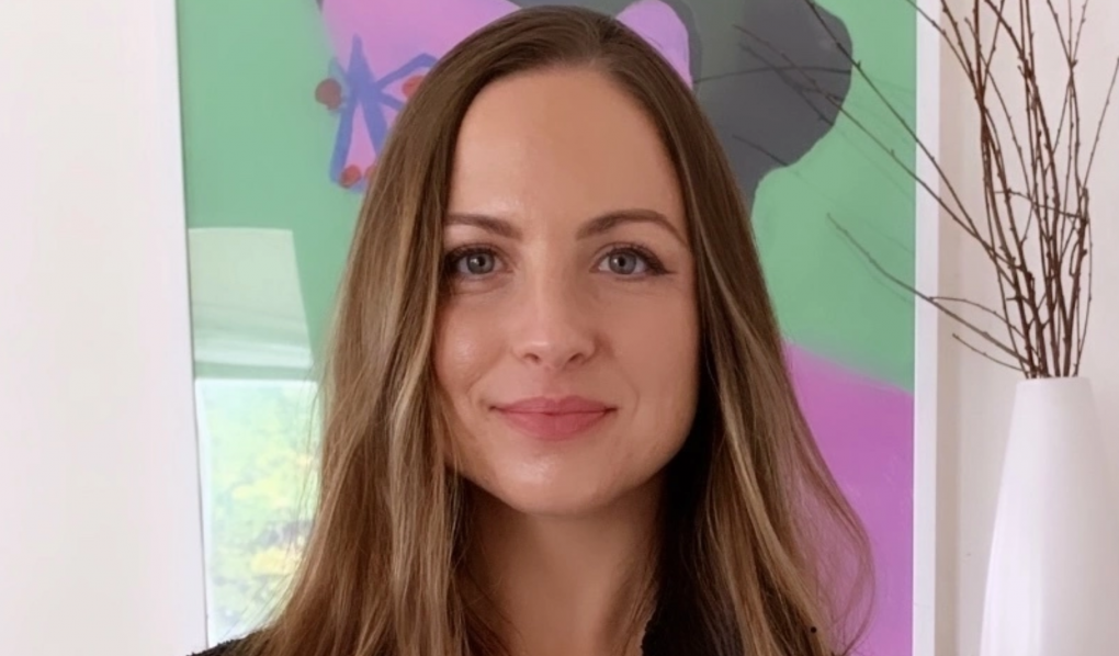 A white woman with long light brown hair faces the camera, half smiling with a colorful painting behind her.