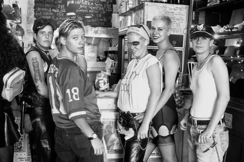 Five butch women with short hair and leather accessories stand at a lunch counter, casually waiting to be served.
