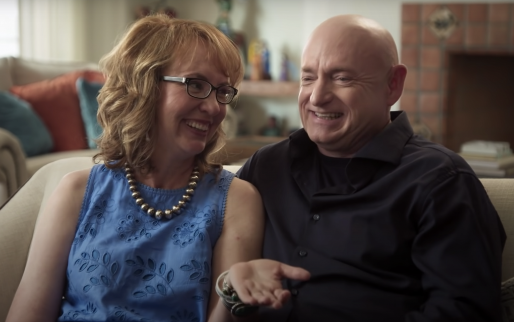 A middle aged woman dressed smartly in a blue dress and statement necklace gestures to her husband, a bald man wearing a black shirt. Both are sitting close together on a couch, laughing.