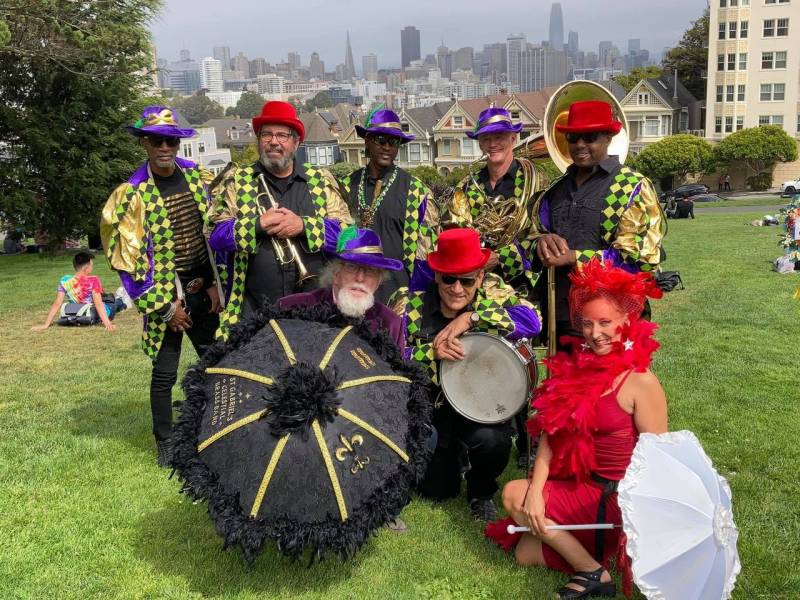 A colorfully dressed eight-person brass band smiles at the camera while posing with drums and bright umbrellas in a San Francisco park.
