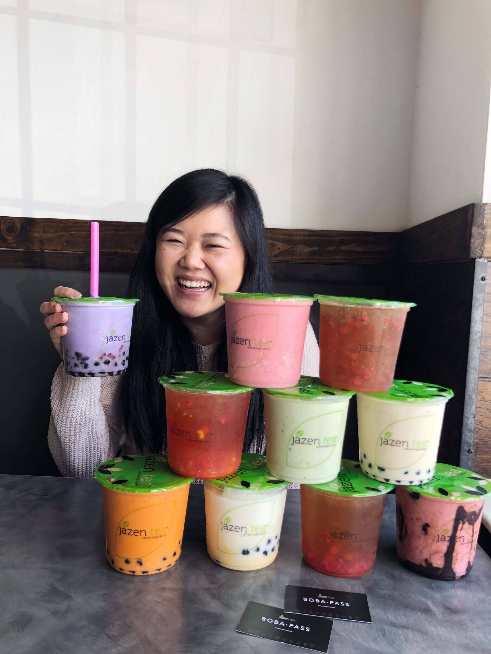 A woman sits in cafe booth with a huge stack of oversized cups of boba in front of her.