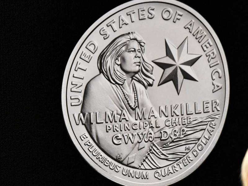 A quarter depicting a windswept Wilma Mankiller wearing a shawl and depicted in side profile.