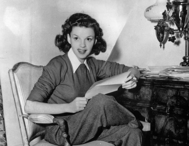 Judy Garland, dressed in loose slacks, a tight sweater with a white collared shirt underneath, sitting forward on a chair next to an ornate wooden desk, papers and a small cigar in hand.