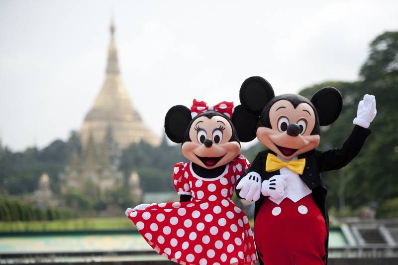 Humans in Mickey and Minnie Mouse costumes stand arm in arm with a temple-like structure behind them in the distance.