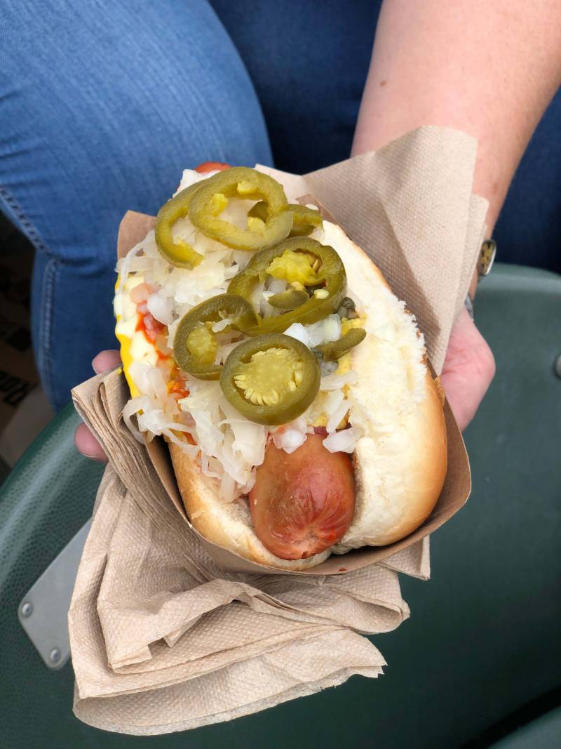 woman holds a hot dog with jalapenos, onions, and ketchup at a baseball game
