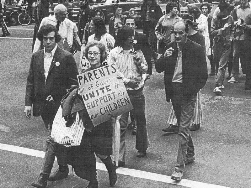  A bespectacled woman dressed in conservative 1970s clothes smiles, as she walks through the street carrying a sign that says "Parents of gays unite in support for our children." Behind her is a group of other men and women walking in formation.