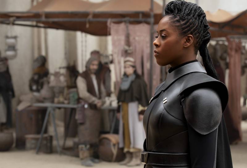 A Black woman with neat braids wears a futuristic black leather uniform and stands to attention in the middle of a marketplace.