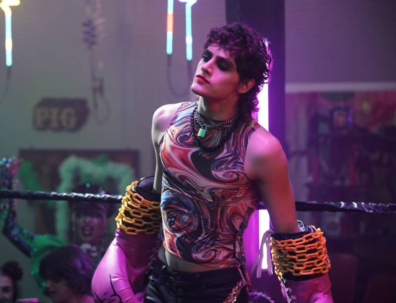 A young man with tousled hair stands boldly. He is wearing dark eye make-up, red lipstick and comedically large purple boxing gloves bound at the wrists with yellow chains.