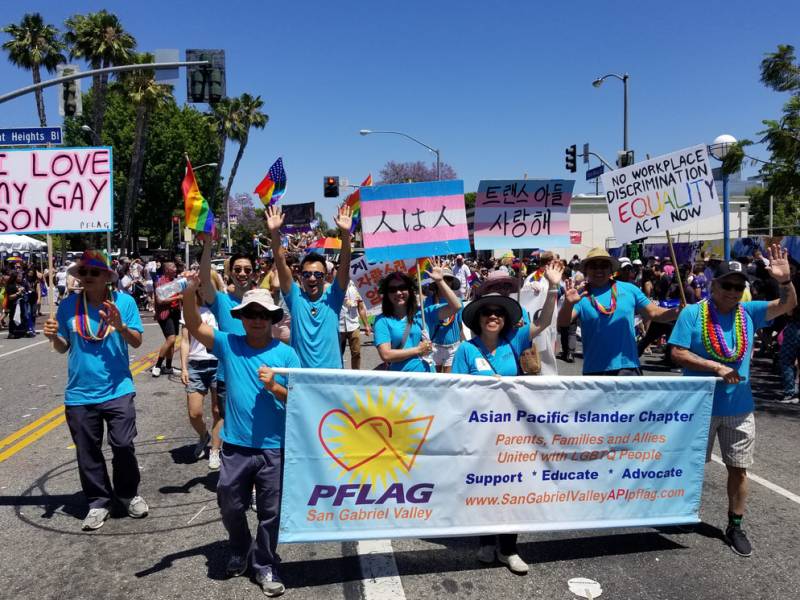 A group of smiling and waving people in matching blue t-shirts lead a celebratory march through sunny streets. Rainbow flags are held aloft in the background.