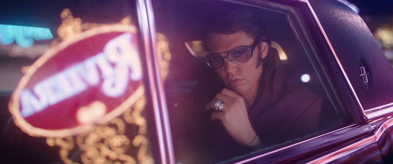 An actor portraying Elvis Presley stares pensively out a limousine window while wearing sunglasses, as the window reflects neon lights outside.