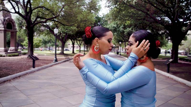two dancers in light blue bodysuits embrace in the middle of a dance on an outdoor plaza