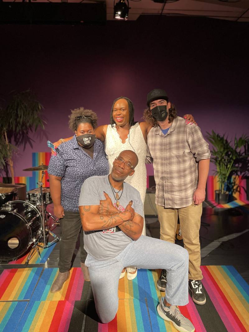 Three artists pose standing up in music performance set with rainbow flag rug, with one person kneeling and crossing arms in front of them. Musical instruments and house plants are present in the background