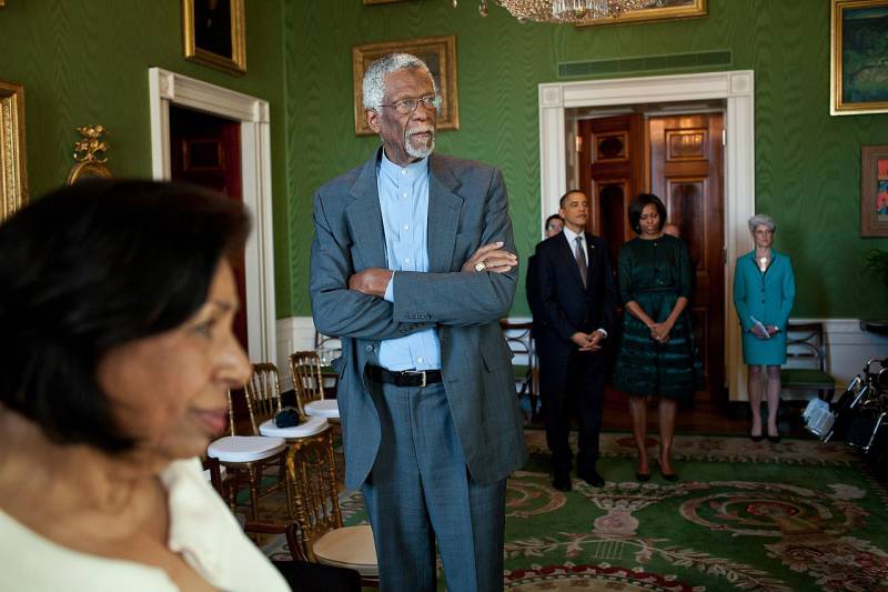 A tall Black man in a blue suit stands in a wing of the White House with the President and First Lady.