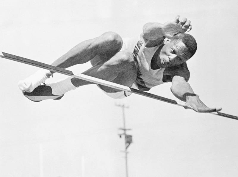 A young Black athlete barely clears a high-jump car against a white background