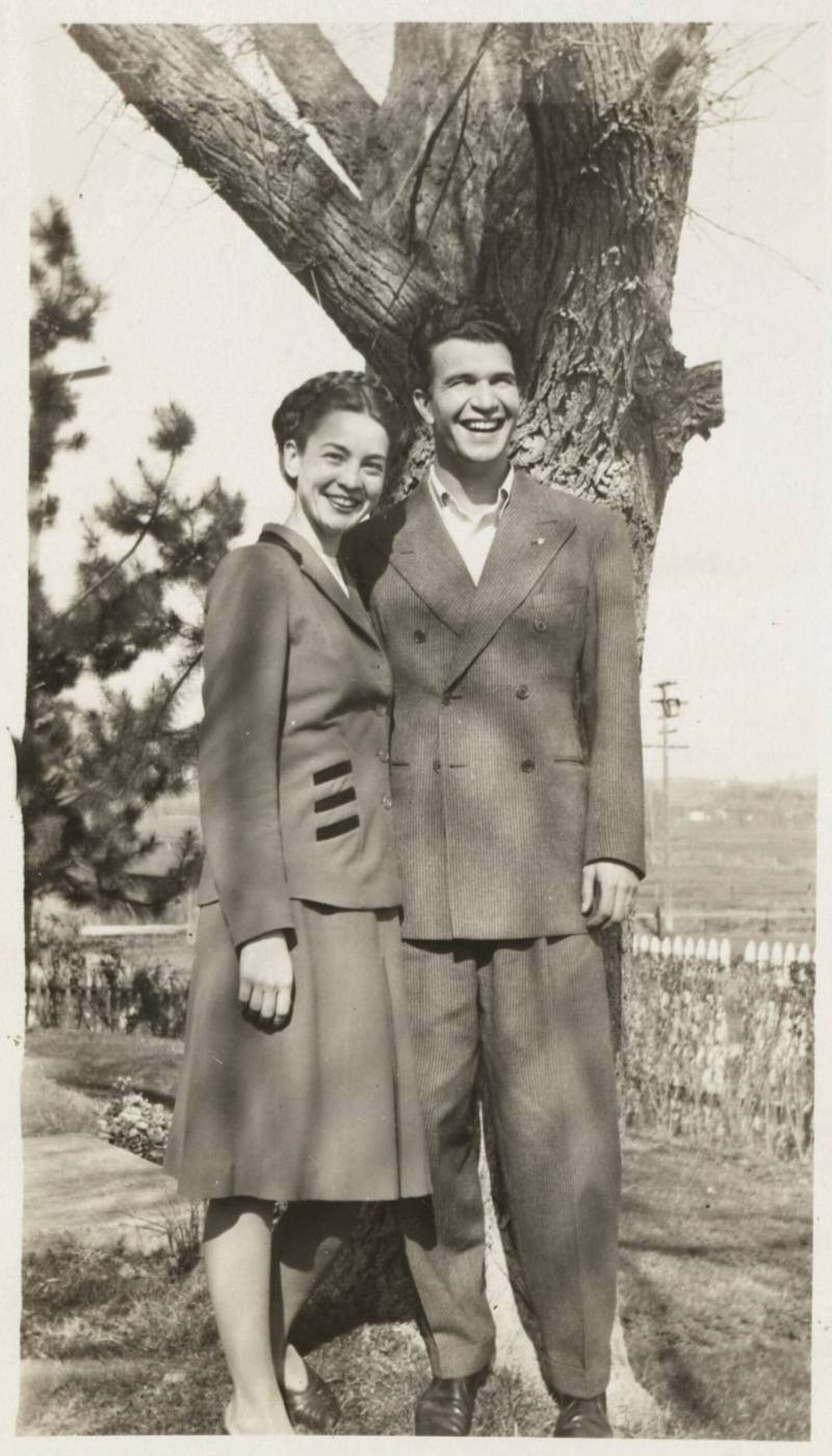 a black and white photo of a man and a woman, smiling with their arms around each other in front of a tree