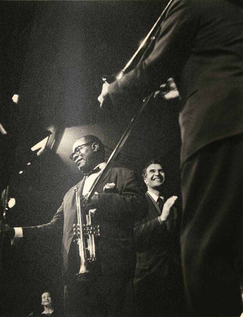 A trumpeter, Louis Armstrong, smiles onstage with another musician, Dave Brubeck, laughing behind him