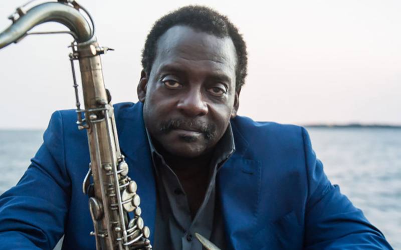 A Black man in a blue suit holds a saxophone while looking into the camera