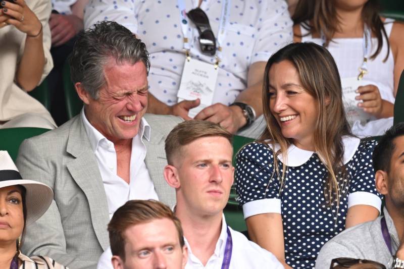 Hugh Grant screws his face up in an enthusiastic laugh alongside a female companion. The pair are sitting behind several rows of serious looking men.