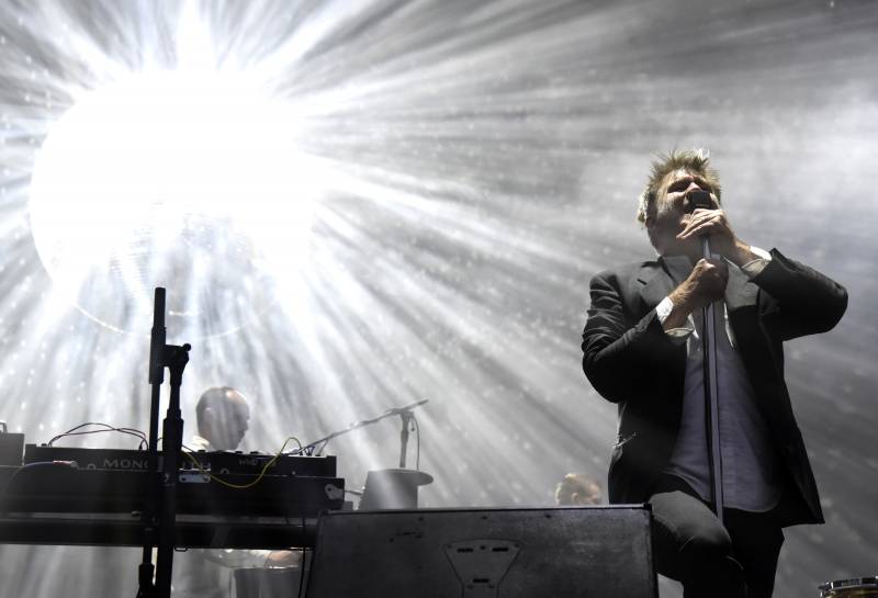a man in a dark suit sings onstage, backlit by a bright light, while a woman plays a keyboard behind him