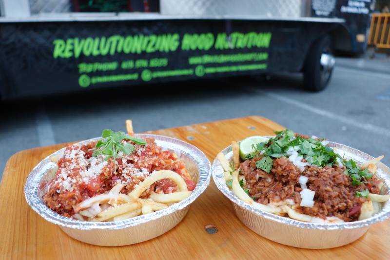 two plates of vegan french fries on a table outside in front of the Vegan Hood Chefs food truck. A printed slogan on the side of the truck says "revolutionizing hood nutrition"